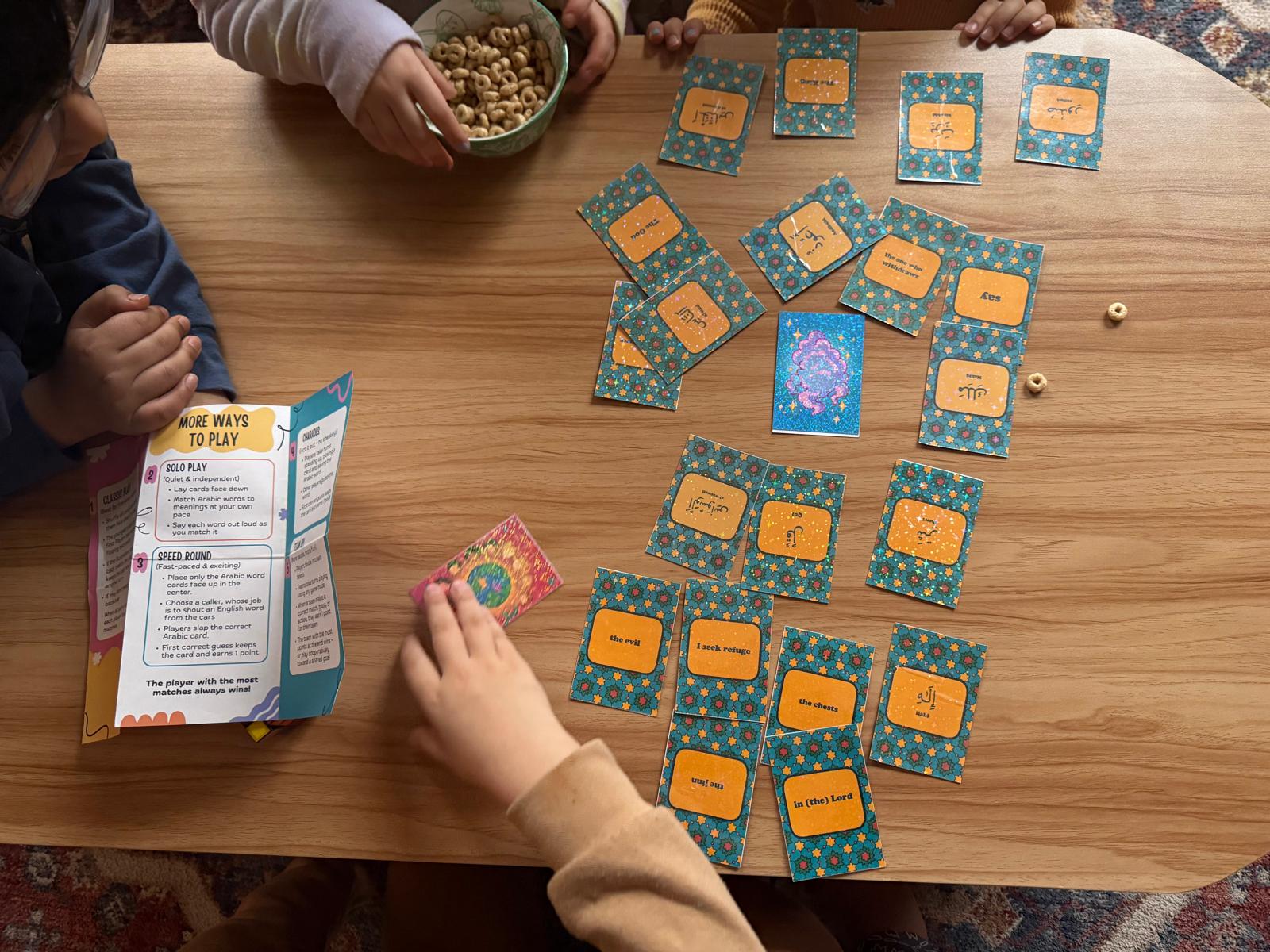 Children playing an colorful Islamic card game to learn Arabic from Qur'an and a bowl of snacks on a wooden table.